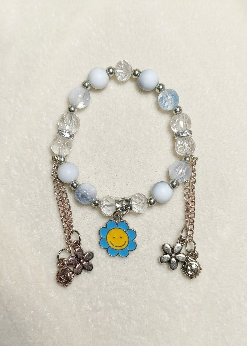A beaded stretch bracelet with white and light blue beads, faceted clear crystal beads, silver-toned spacers, and three dangling charms: a blue flower with a yellow smiley face, a rose-gold-toned flower, and a silver-toned flower.