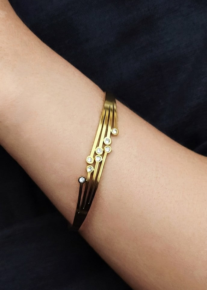 A gold-toned multi-strand cuff bracelet with small clear crystal accents, shown worn on a person's wrist against a dark navy fabric background.