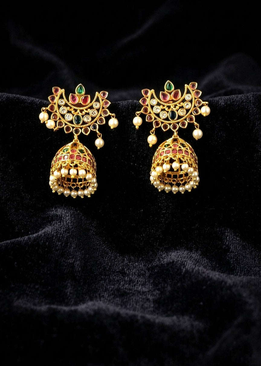 A pair of ornate gold-toned jhumka earrings with red and green enamel details, set against a black velvet background.