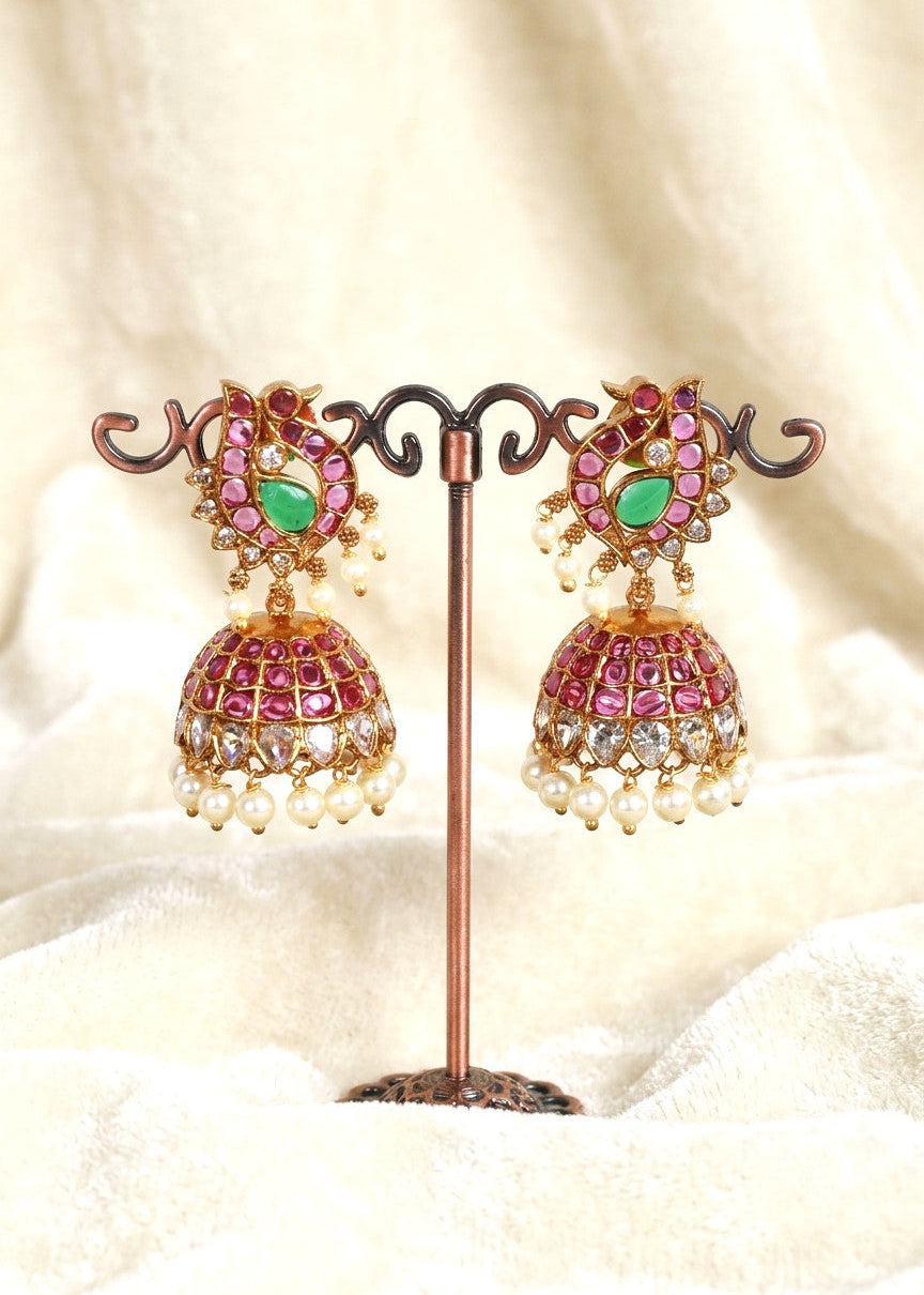 A pair of ornate gold-toned jhumka earrings with pink and green stones and white pearl bead drops, displayed on a decorative stand against a soft cream fabric background.