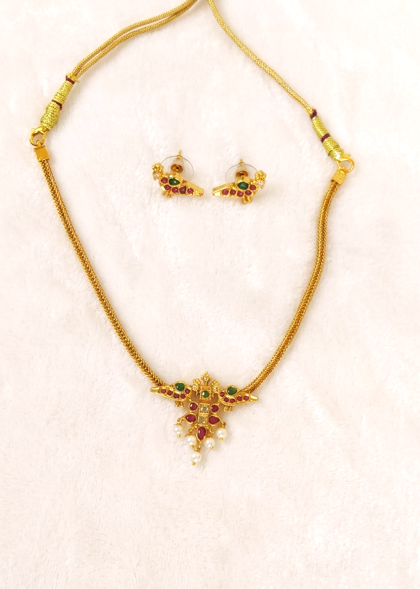 Gold-toned necklace and matching stud earrings set with small green and red stones and white pearl-like accents, displayed on a light pink textured background.