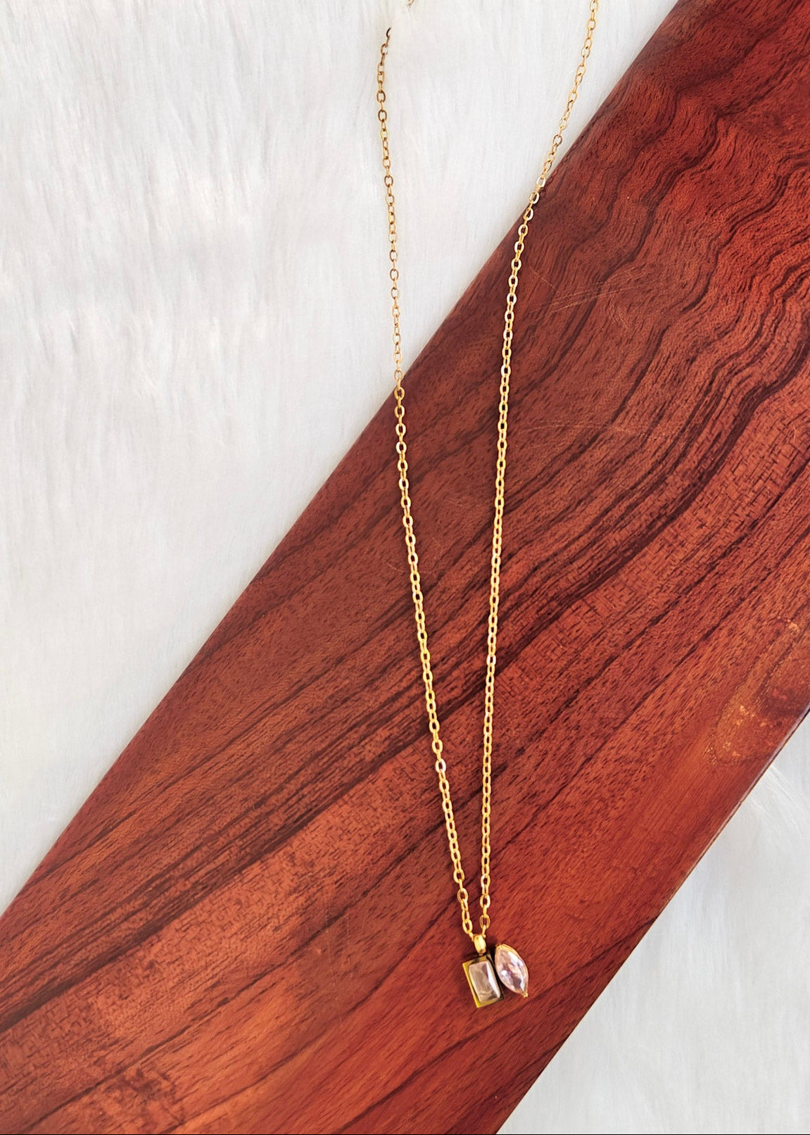 A delicate gold chain necklace with a small pendant featuring a pink stone, displayed on a diagonal wooden block over a white textured background.