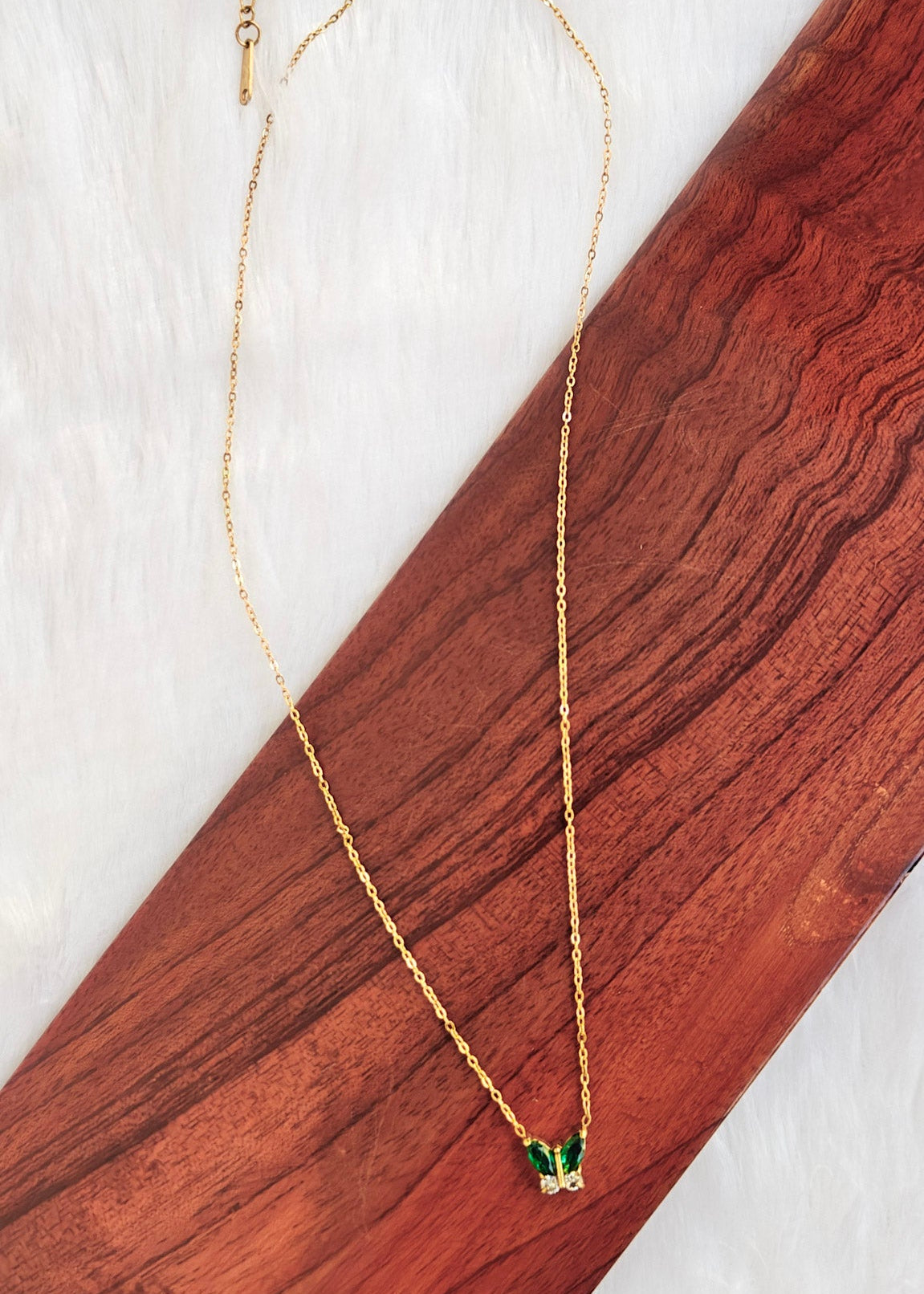 A delicate gold chain necklace with a small butterfly pendant featuring green wings and clear stones, displayed on a diagonal wooden block over a white textured background.