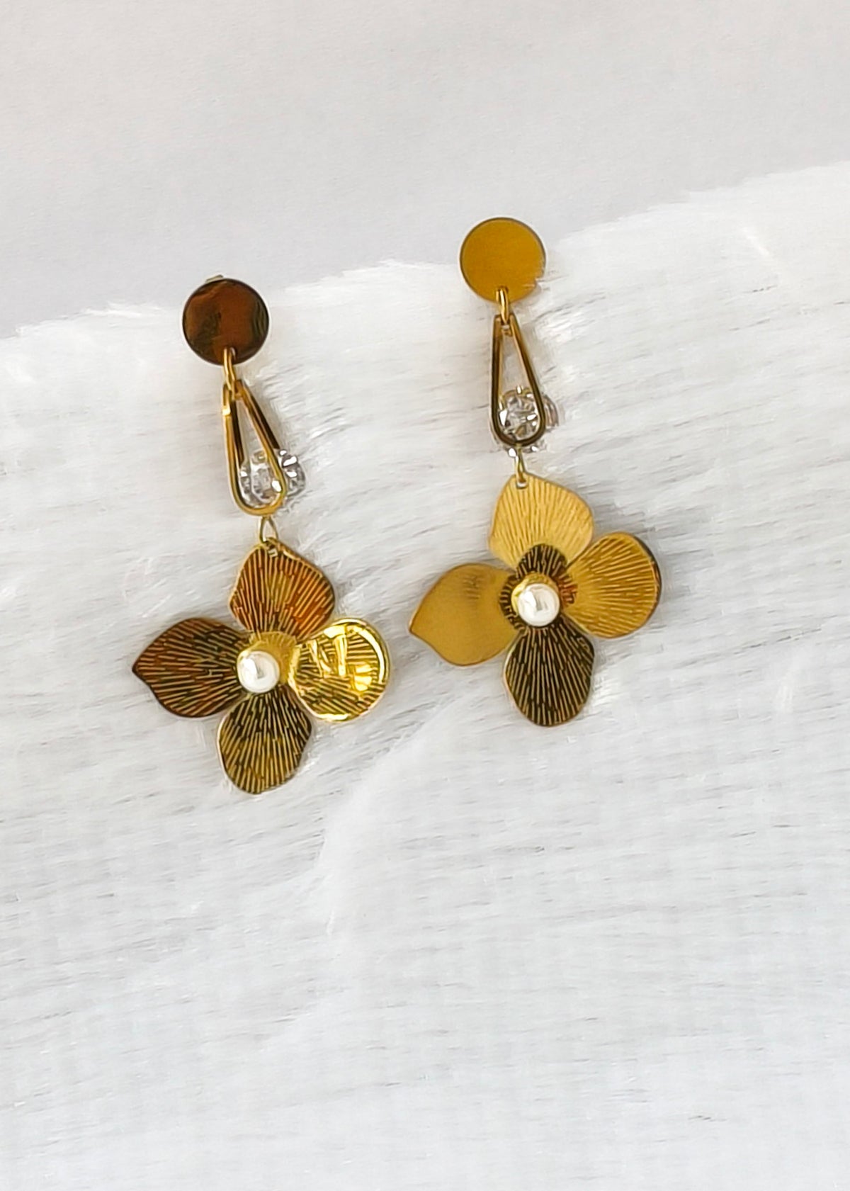 A pair of gold and bronze flower-shaped drop earrings with textured petals, clear rhinestone accents, and small pearl-like centers, displayed on a white textured fabric background.