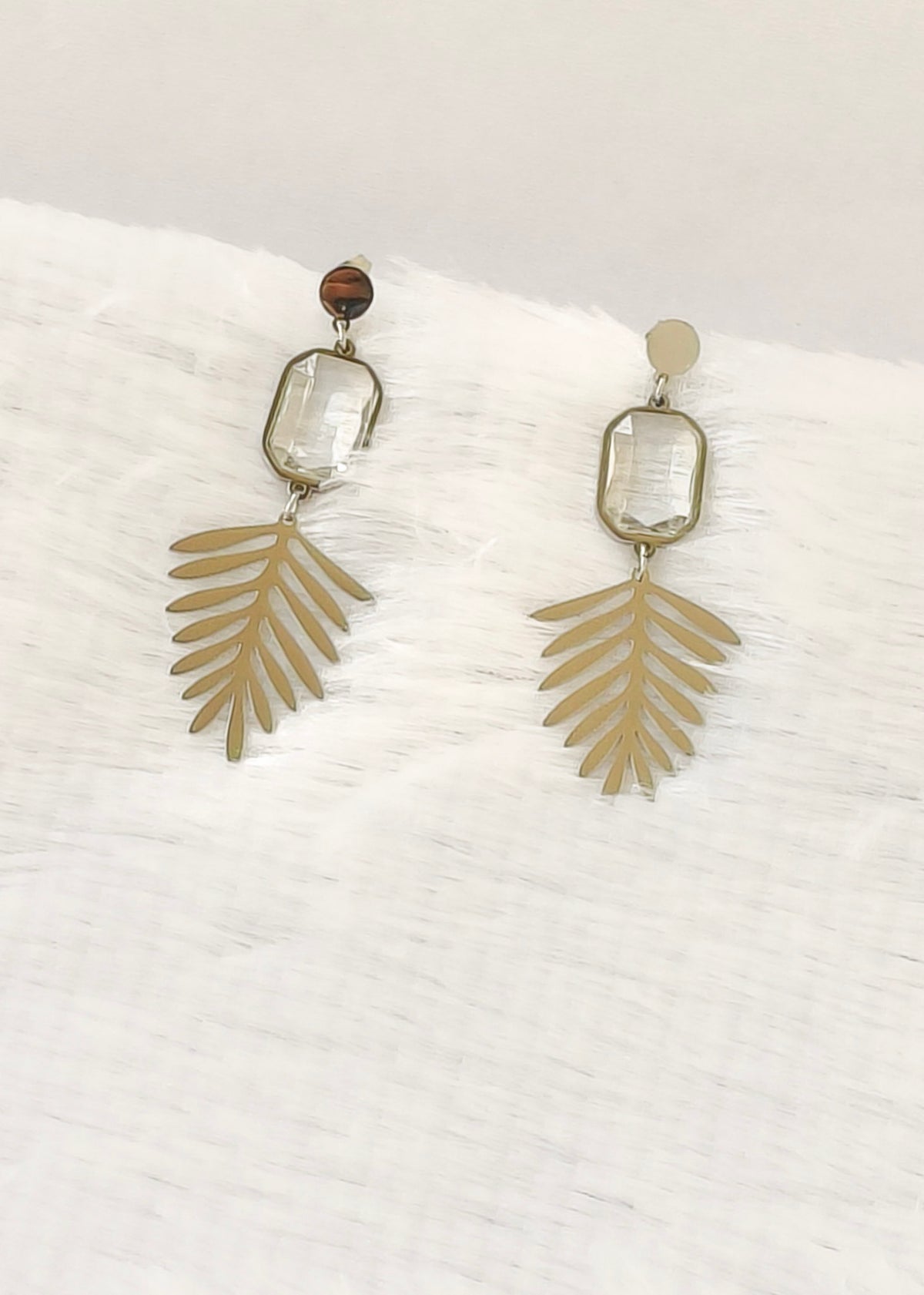 A pair of bohemian-style dangle earrings featuring gold-toned palm leaf charms, clear rectangular glass beads, and a small brown tiger's eye bead, displayed on a white textured fabric background.