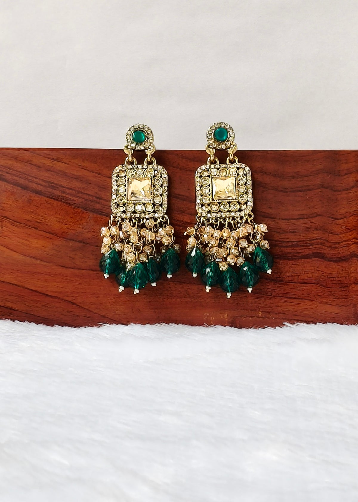 A pair of ornate gold-toned chandelier earrings with green stones and clear crystal-like accents, displayed on a wooden block against a white background.