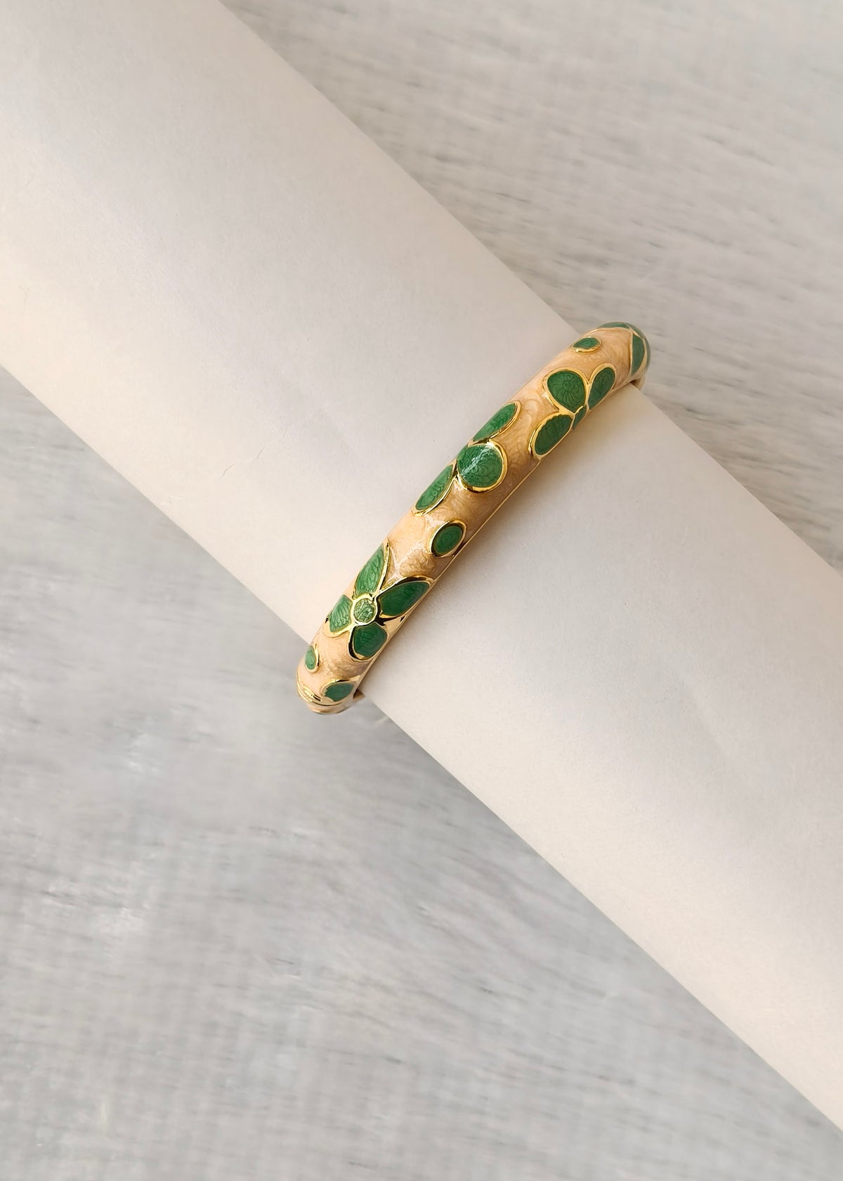 A gold bangle bracelet with green enamel leaf-like inlays, displayed on a white cylindrical stand against a light gray textured background.