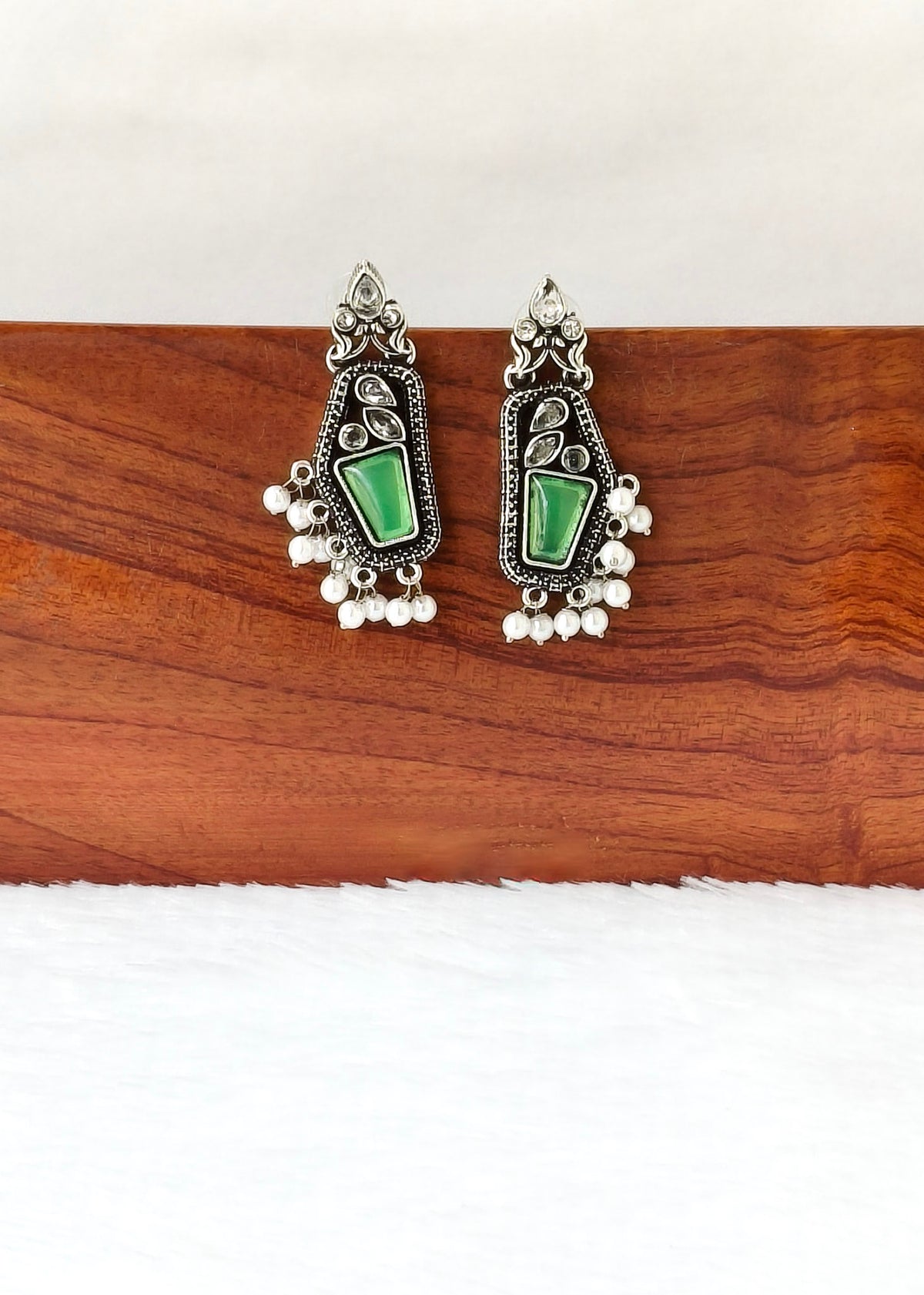 A pair of silver-toned traditional Indian earrings with green stone inlays and dangling pearl-like beads, displayed on a wooden block against a white background.