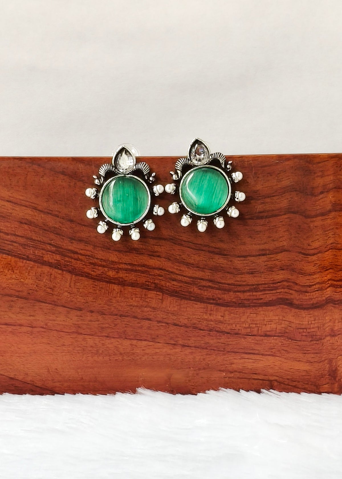 A pair of silver-toned statement earrings with oval green stones and small clear crystal-like accents, displayed on a wooden block against a white background.