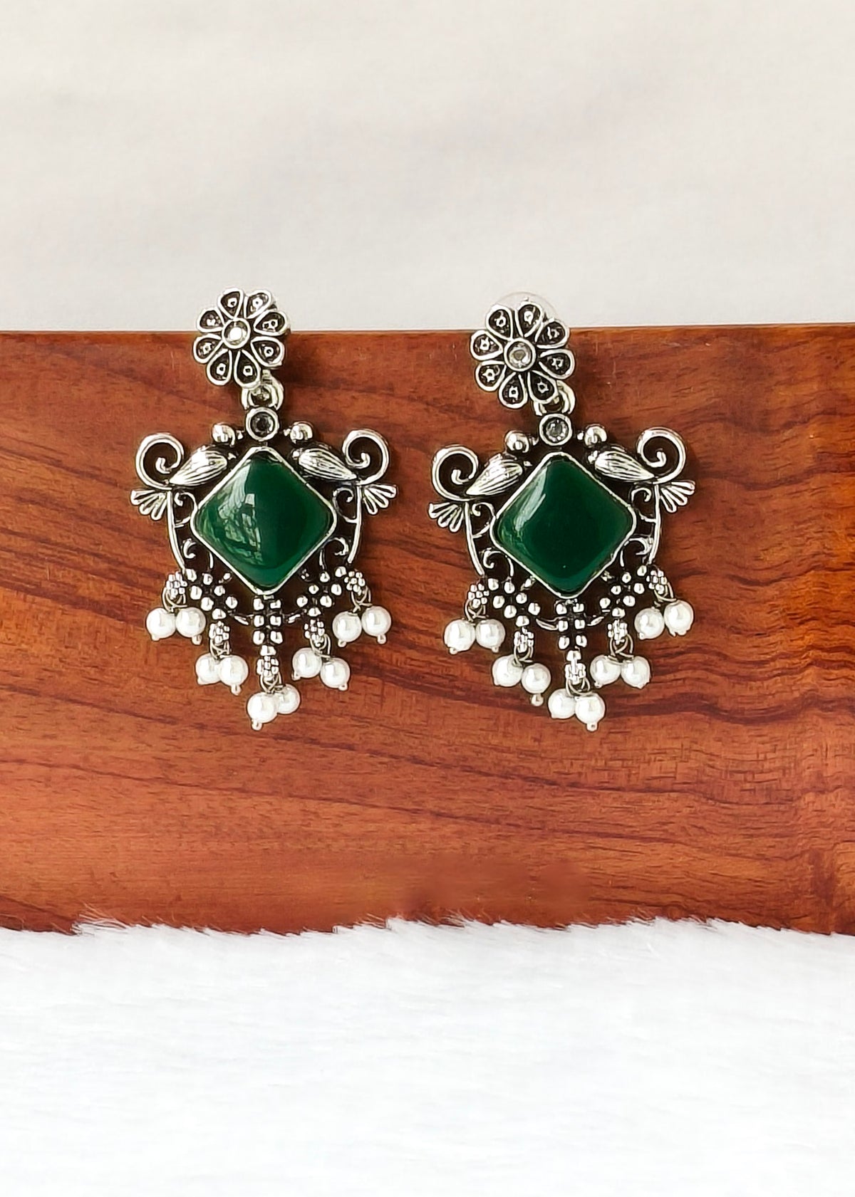 A pair of ornate silver-toned earrings with green square stones and dangling pearl-like beads, displayed on a wooden block against a white background.