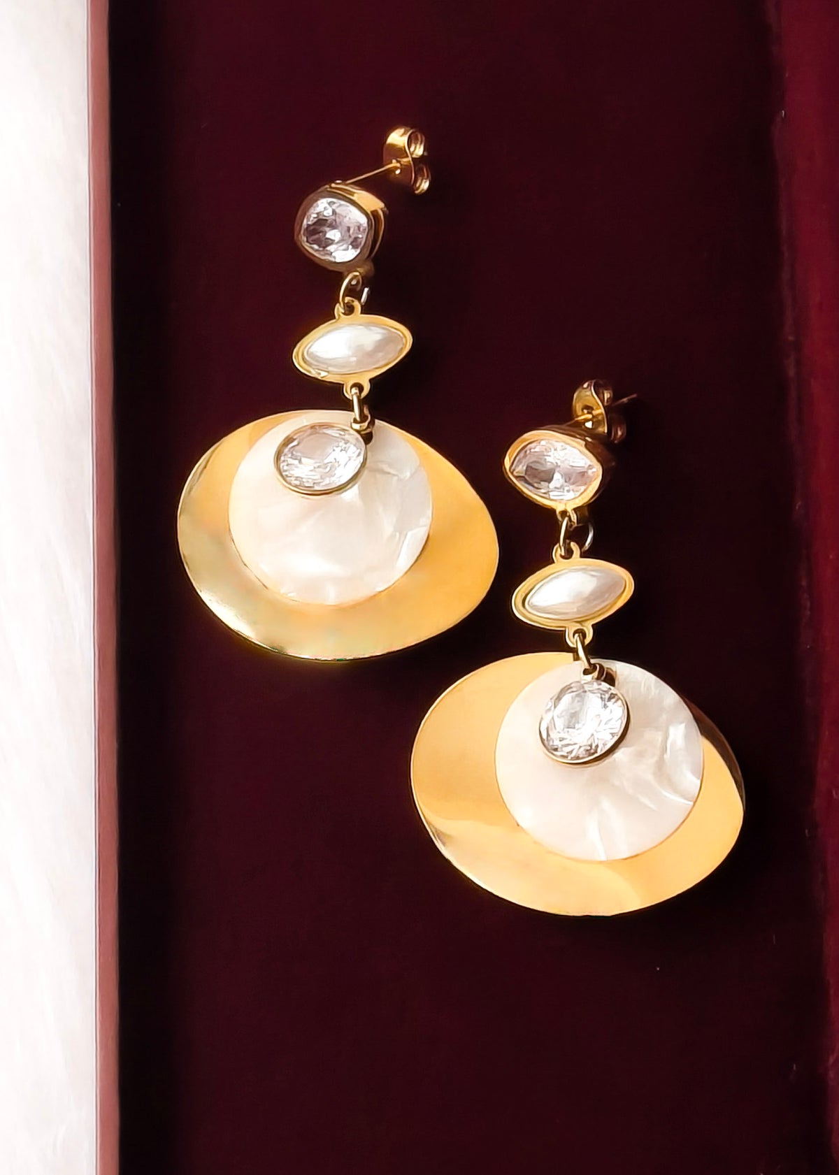 A pair of gold drop earrings with large gold disc-shaped elements, white mother-of-pearl oval discs, and clear rhinestone accents, displayed on a dark burgundy velvet surface.