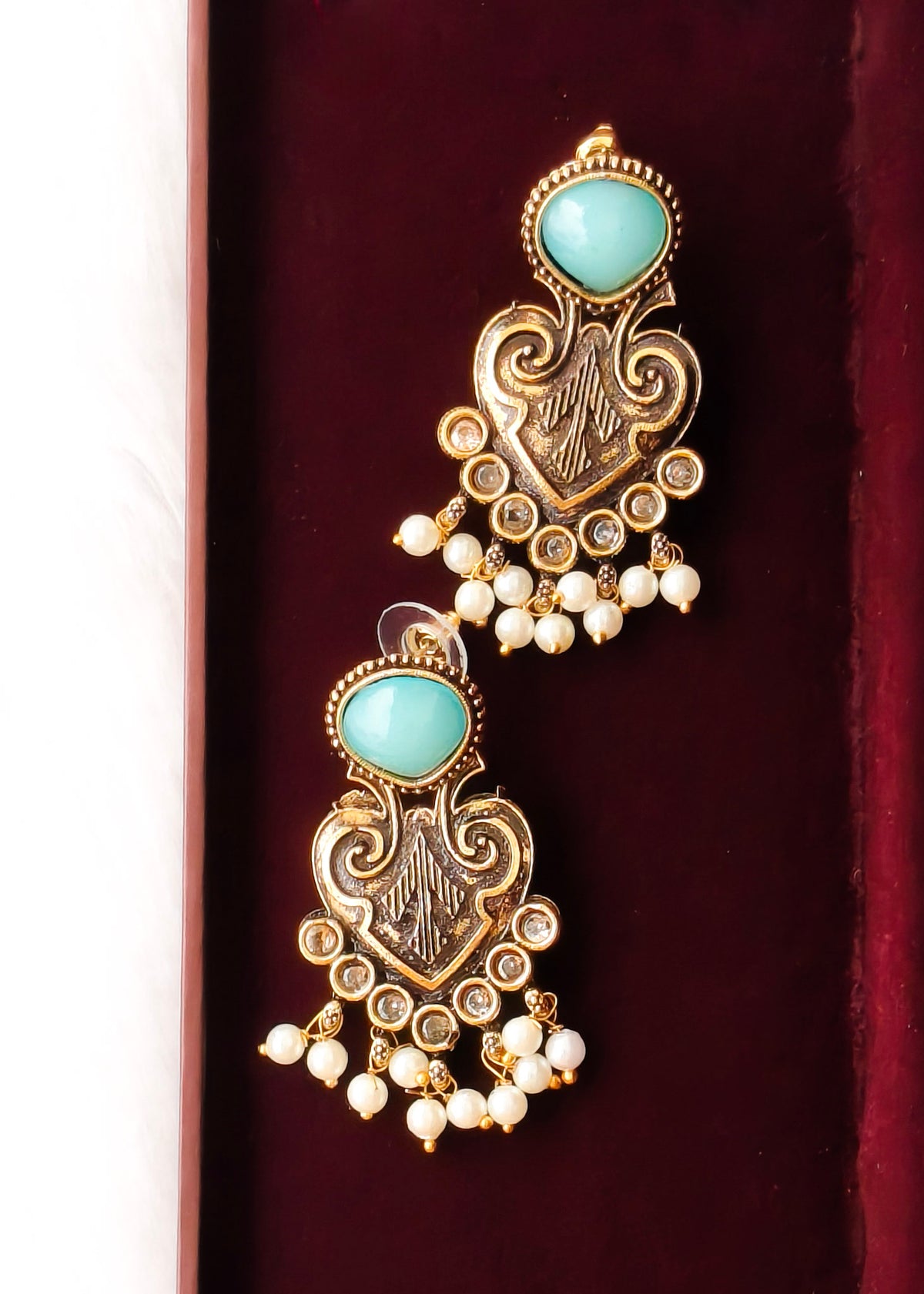 A pair of ornate gold-toned dangle earrings with oval light blue-green stones and small white pearl-like drops, displayed on a dark burgundy velvet surface.