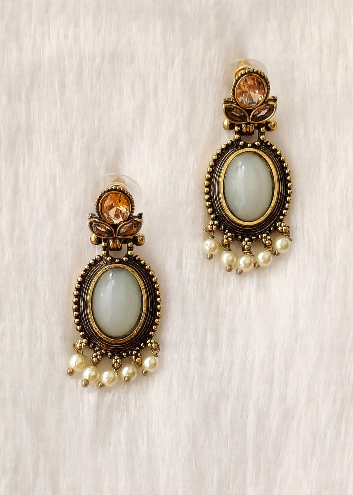 A pair of antique bronze drop earrings with oval light green stone centers, small amber crystal accents at the top, and dangling pearl beads at the bottom, displayed on a soft white textured fabric background.