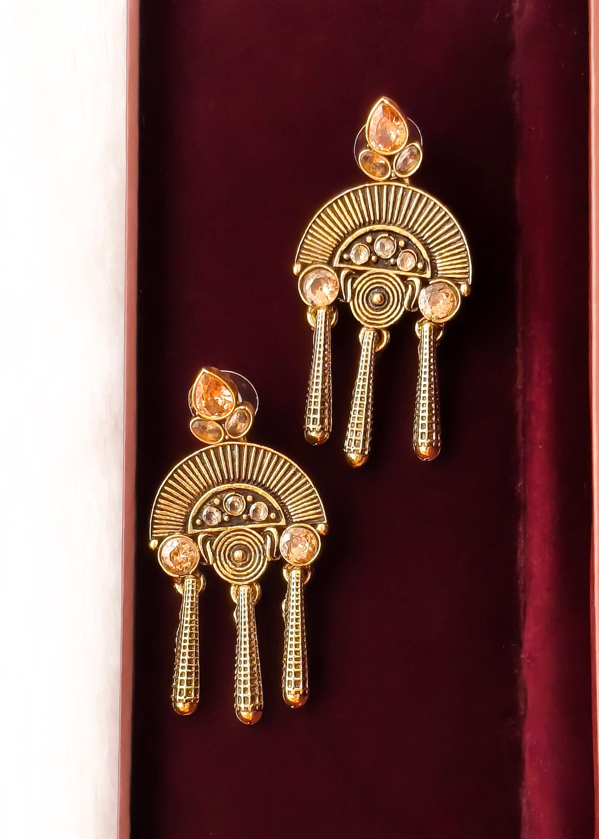 A pair of ornate gold-toned traditional earrings with circular fan-like motifs, small clear stones, and dangling bead-like elements, displayed on a deep burgundy velvet surface.
