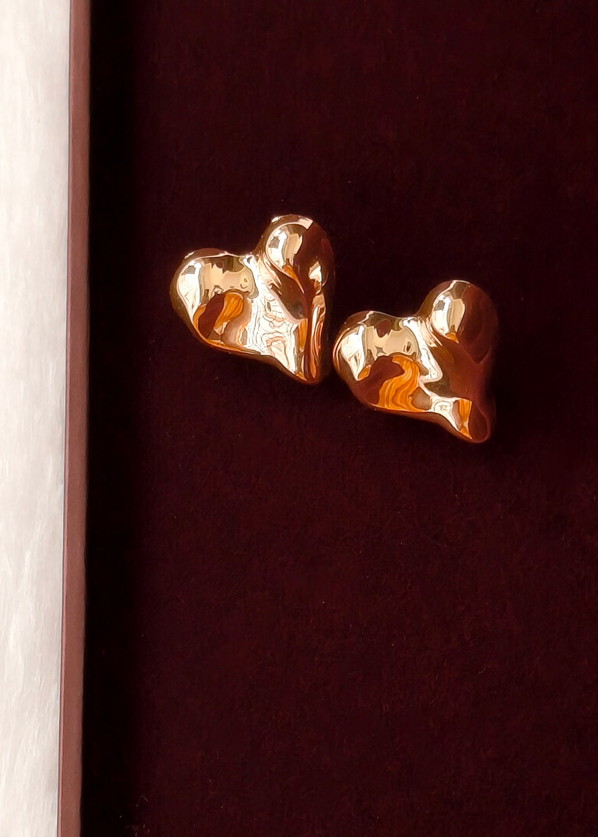 A pair of gold-toned, textured heart-shaped stud earrings displayed on a dark burgundy velvet jewelry box, with a light wood sliver visible on the left edge.