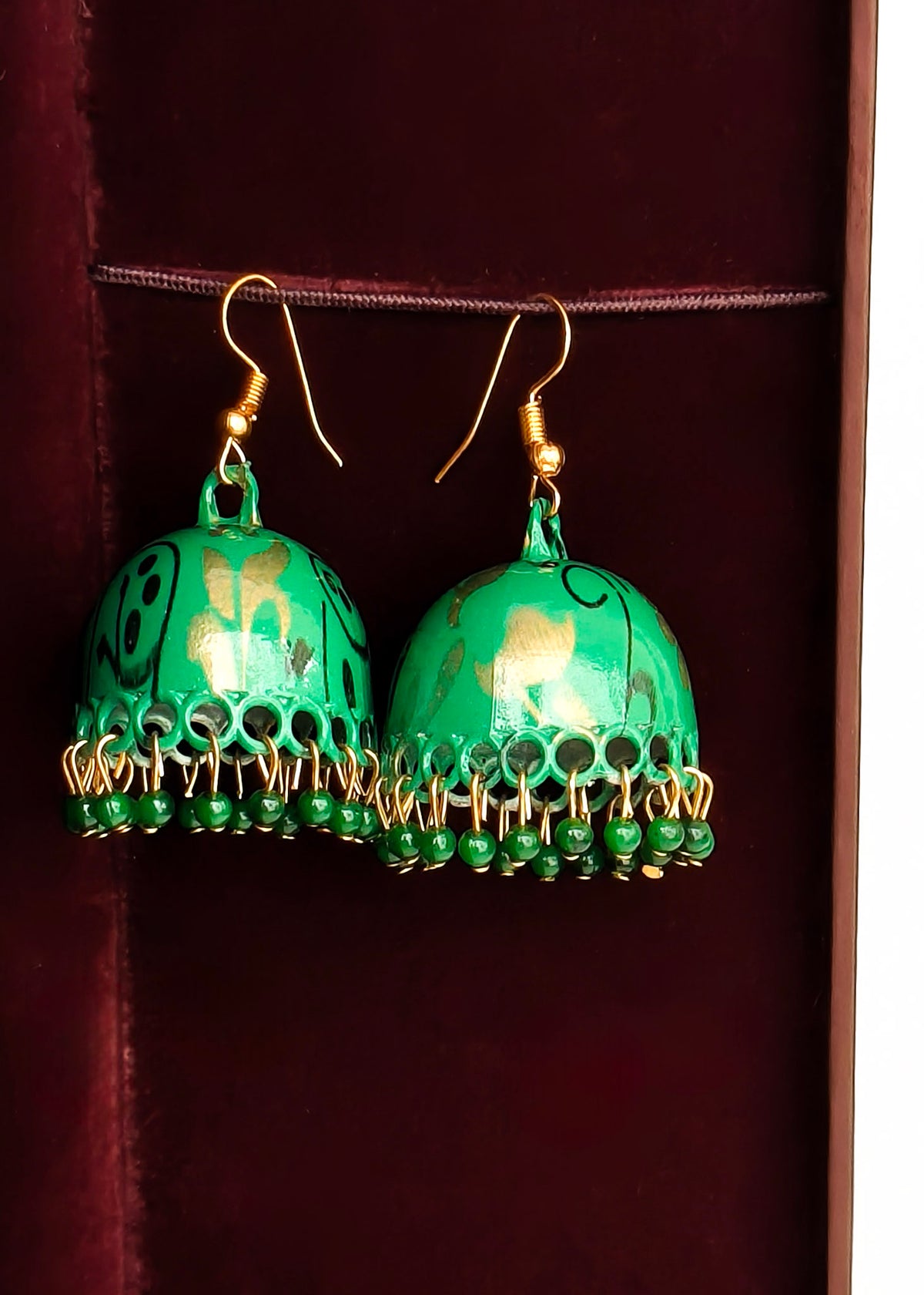 A pair of green jhumka earrings with gold-tone hooks and dangling beaded fringe, displayed against a dark red velvet jewelry box interior.