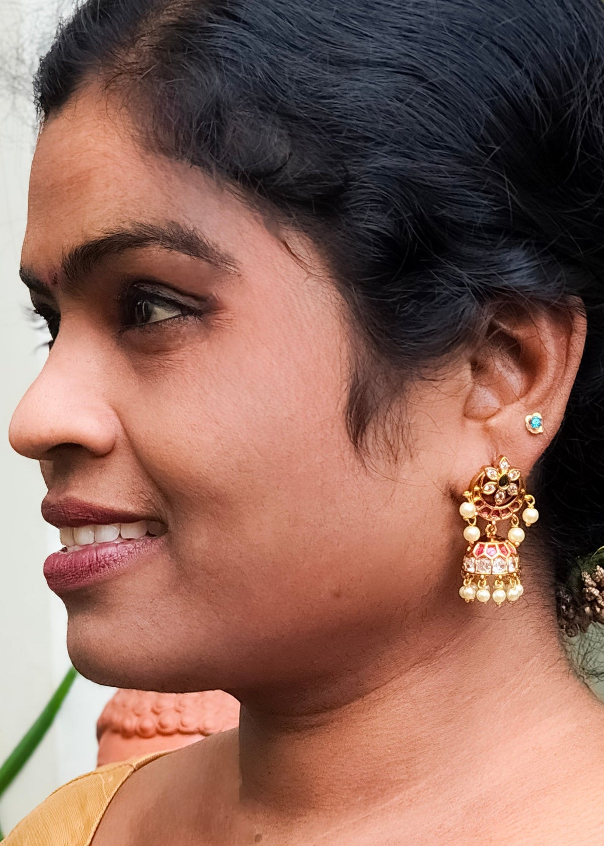 A close-up side profile of a woman wearing a traditional Indian gold Jhumka earring with a small green stud earring.