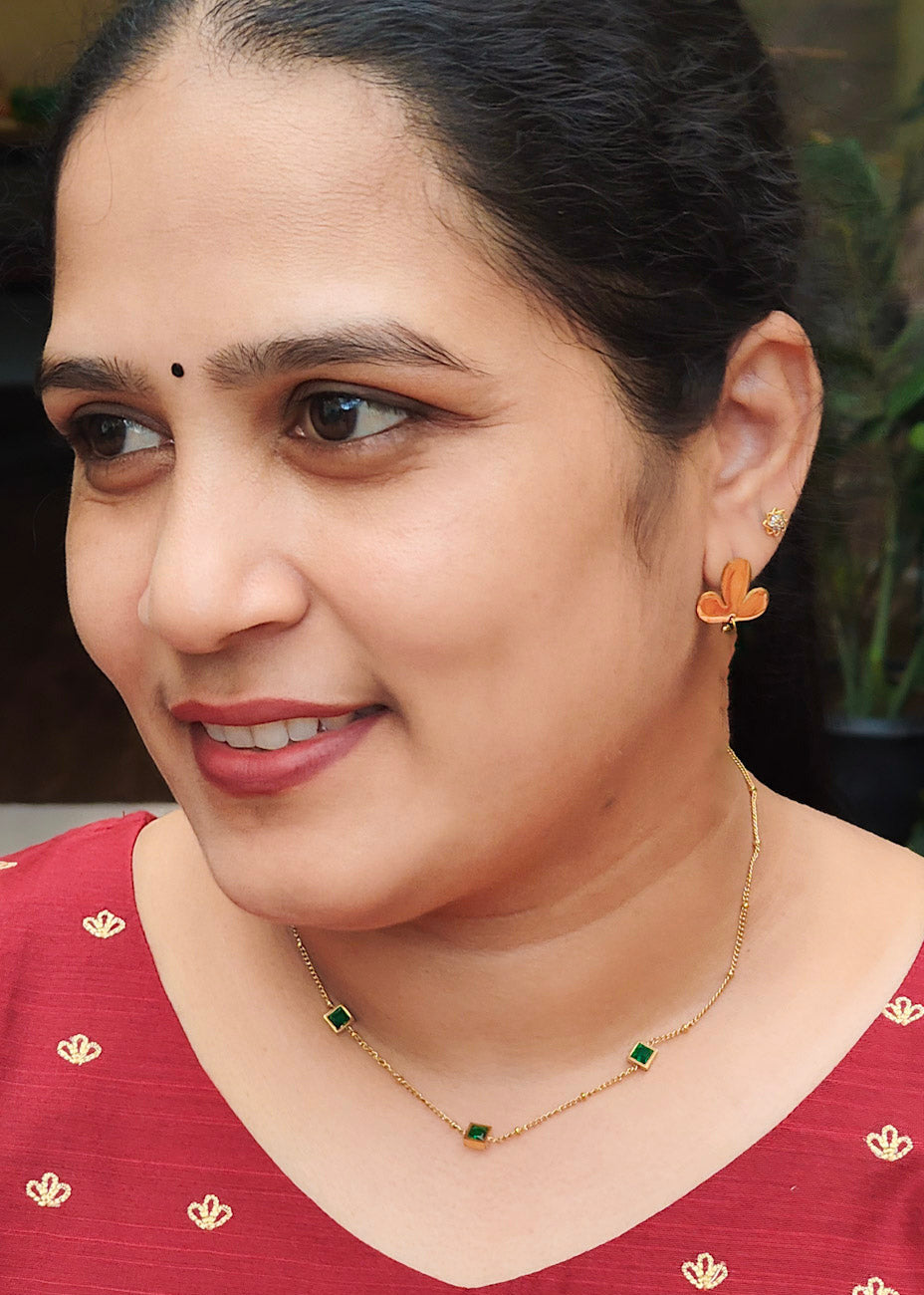 A woman with dark hair pulled back, wearing a red top with gold floral embroidery and a gold necklace with green stones, shown in a close-up portrait.