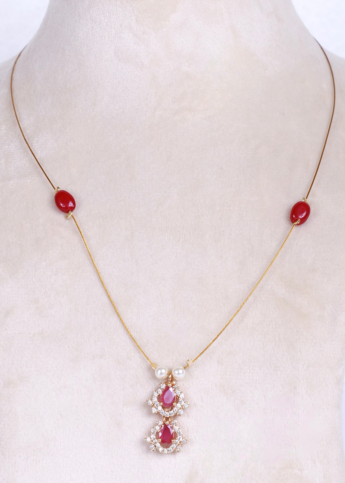 A gold chain necklace displayed on a cream-colored mannequin bust, featuring two small red oval beads on the chain and a central pendant with red teardrop-shaped stones surrounded by clear crystals.