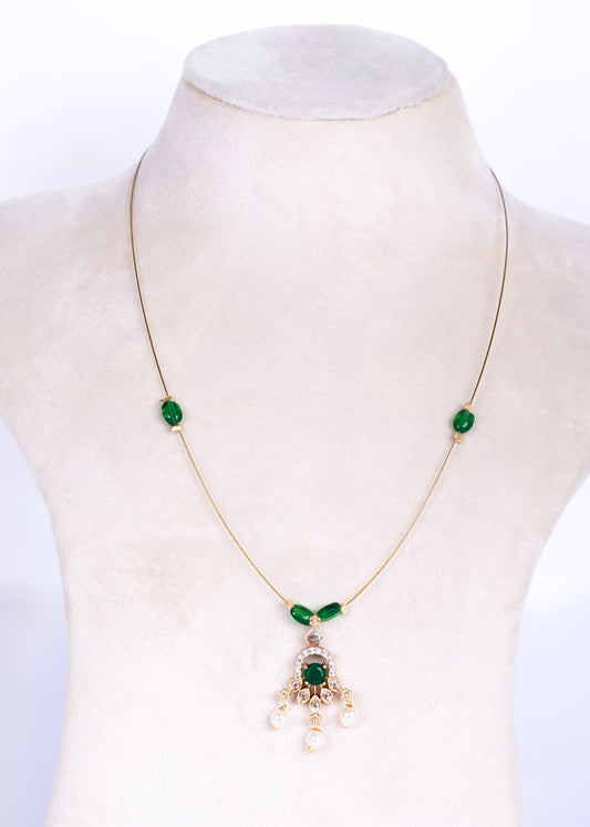A gold chain necklace displayed on a cream-colored jewelry bust. The necklace features two small green oval stones on either side of the chest, a small green leaf-shaped accent at the center of the chain, and a central pendant with a larger green oval stone, gold detailing, and three dangling white pearl-like beads.