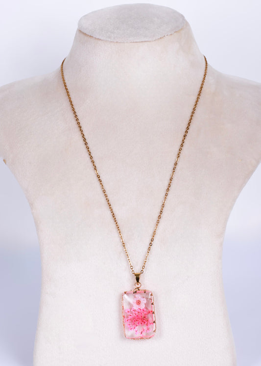A rectangular pendant necklace with a pink and white floral pattern, suspended on anti tarnish gold-toned chain, displayed on a cream-colored mannequin bust.