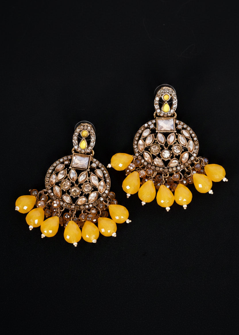 A pair of ornate Indian jhumka earrings with gold-toned metalwork, clear stone embellishments, and dangling yellow teardrop beads.
