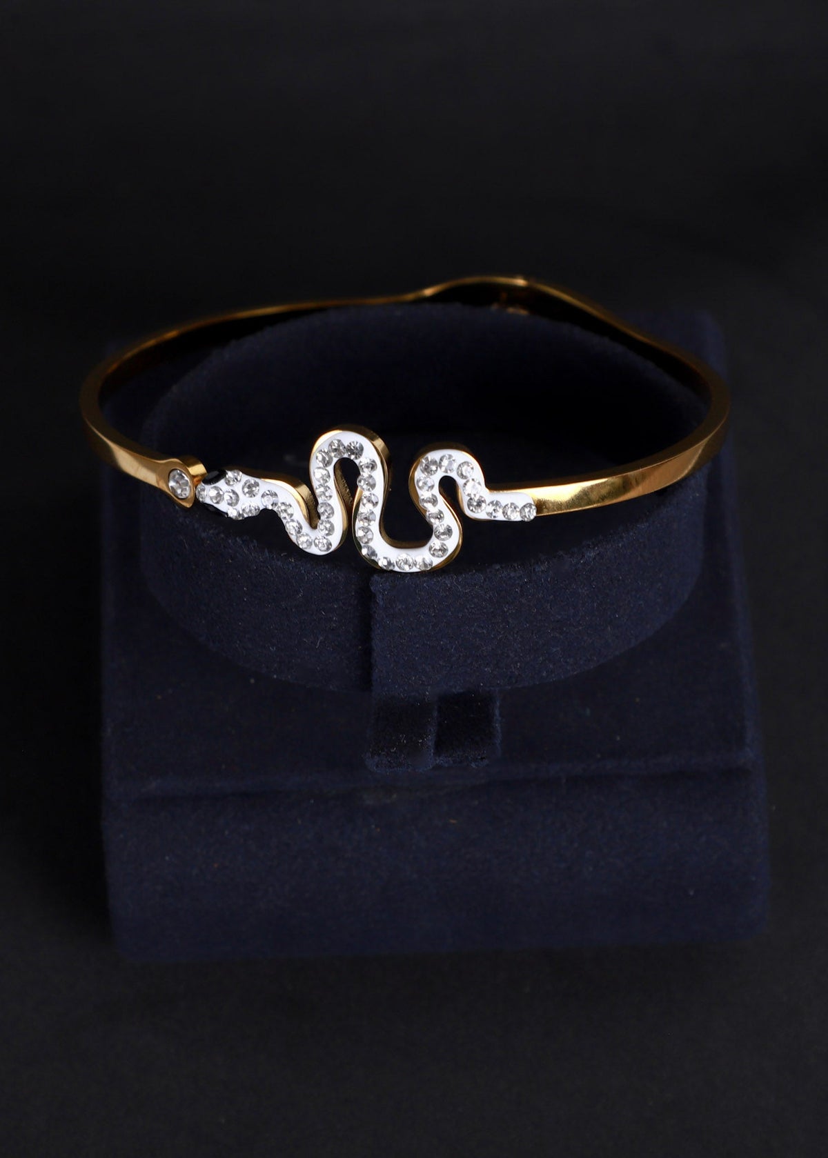 A gold snake-shaped cuff bracelet adorned with clear rhinestones, displayed on a dark navy velvet jewelry box against a black background.