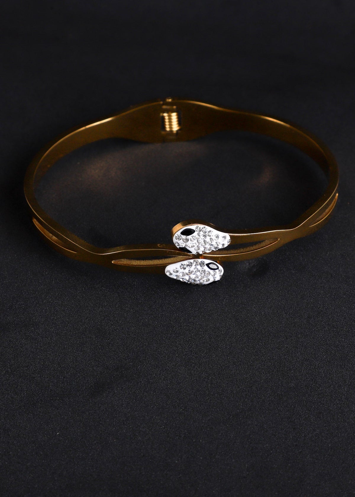 A gold-plated bangle bracelet with two small fish charms encrusted with clear rhinestones, displayed on a black textured background.
