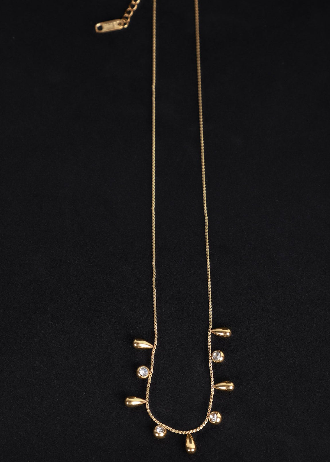 A gold chain necklace with small gold teardrop charms and clear rhinestone accents, displayed against a solid black background.