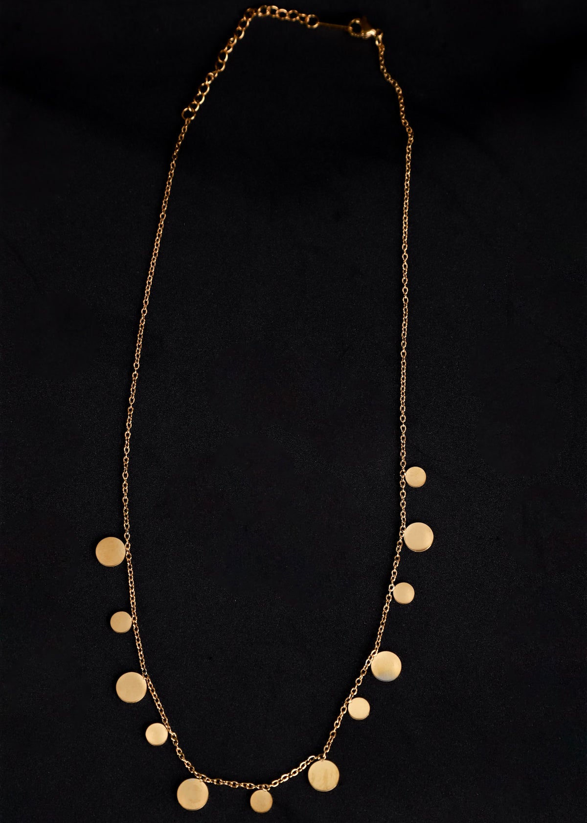 A gold chain necklace featuring multiple flat, disc-shaped metal pendants of varying sizes, displayed against a solid black background.