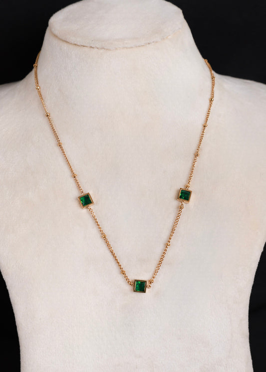 A gold anti-tarnish chain necklace with three small square green gemstones, displayed on a cream-colored jewelry bust against a black background.