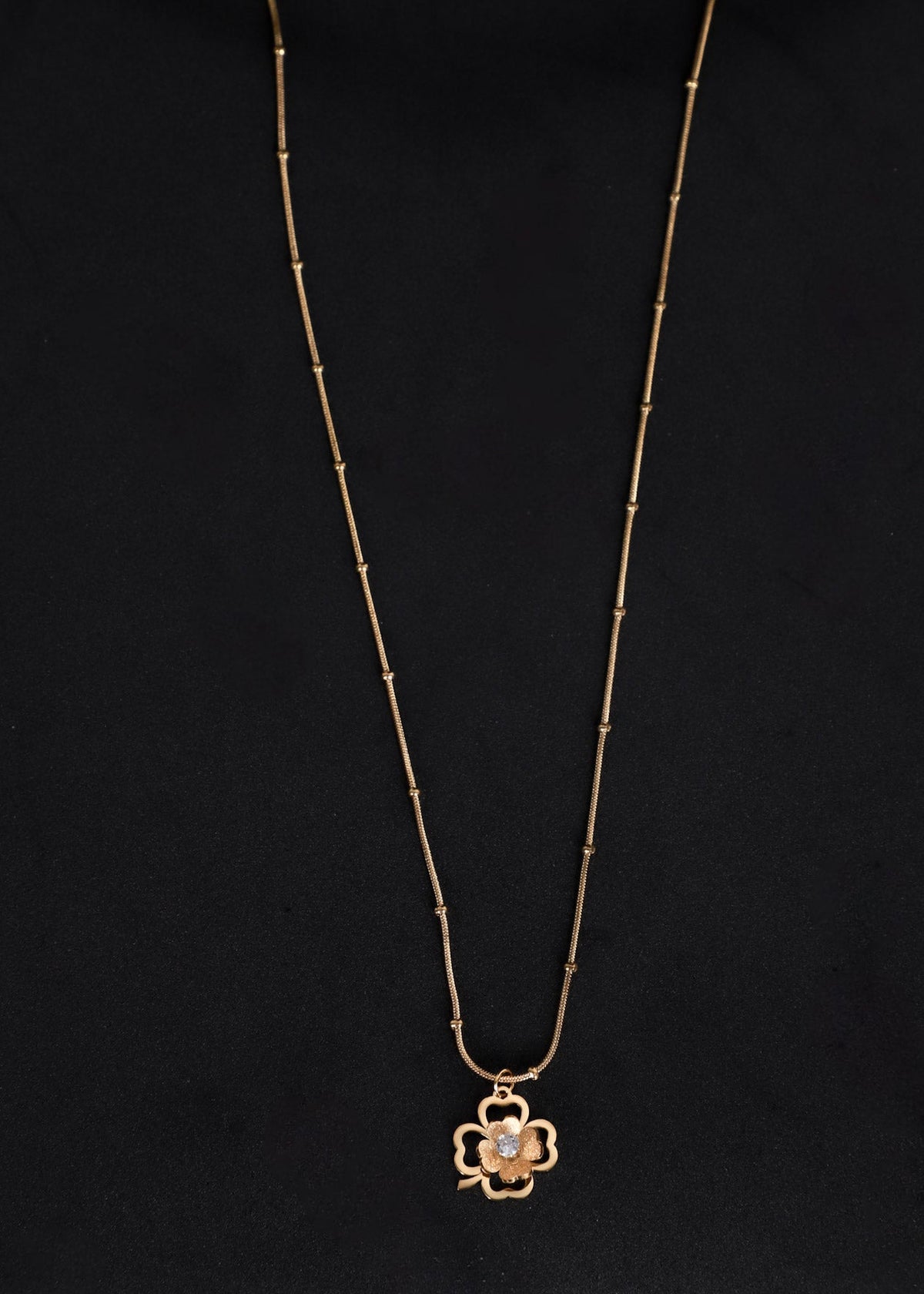 A delicate gold chain necklace with a small clover-shaped pendant featuring a central clear stone, displayed against a solid black background.