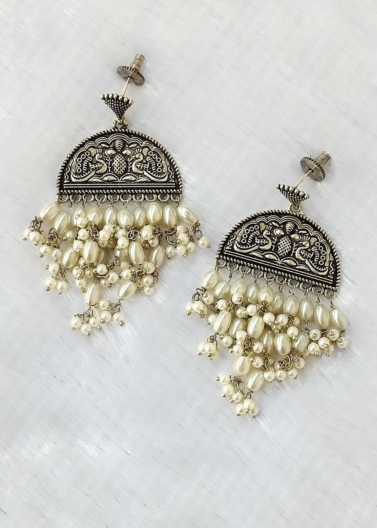 A pair of ornate silver-toned dangle earrings with intricate filigree detailing and cascading cream-colored pearl-like beads, displayed on a textured white surface.