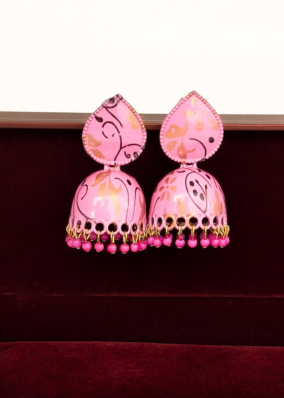 A pair of pink jhumka earrings with gold and black painted patterns, hanging from gold-toned chains with dangling pink beads, displayed against a dark red velvet surface.