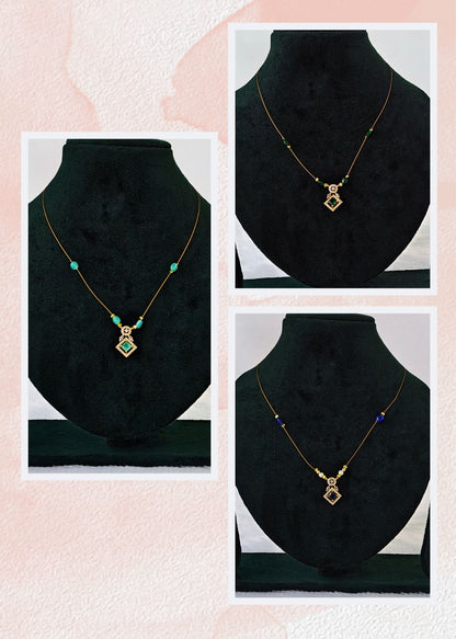 Three invisible gold necklace displayed on black velvet jewelry busts against a light pink textured background. The necklace features a geometric pendant with green and gold details, and small turquoise and purple beads on the chain.
