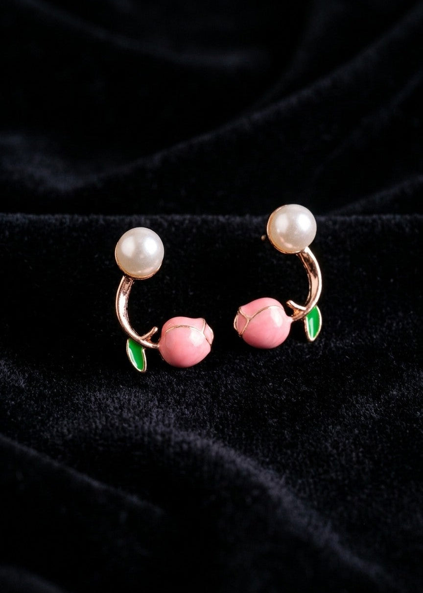 A pair of peach-shaped hoop earrings with pink peach charms, green leaves, and white pearl-like tops, displayed on a black velvet fabric background.