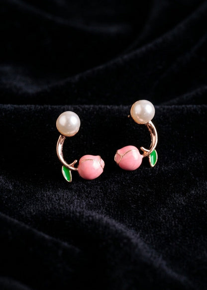 A pair of peach-shaped hoop earrings with pink peach charms, green leaves, and white pearl-like tops, displayed on a black velvet fabric background.