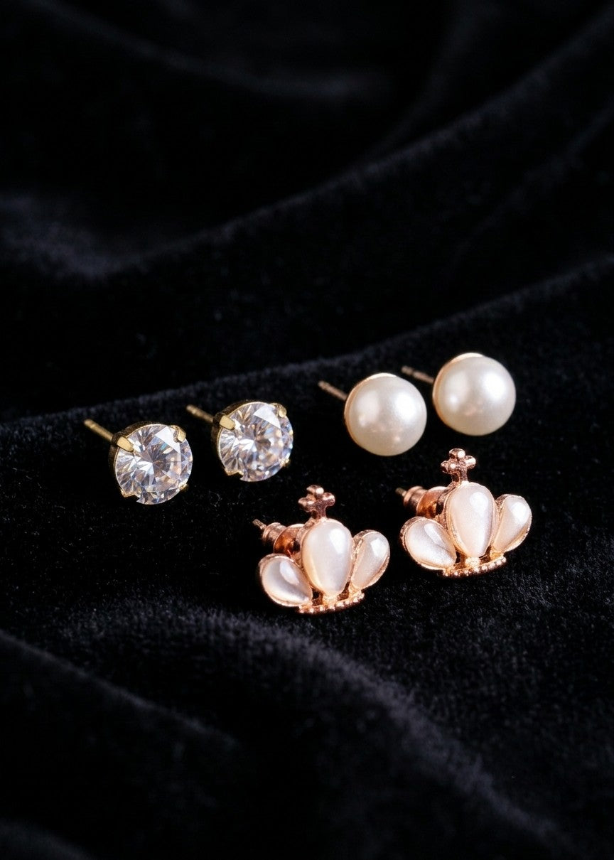 Three pairs of stud earrings displayed on a black velvet background: two pairs with clear round gemstones and one pair with white pearl-like inlays in a rose gold crown setting.