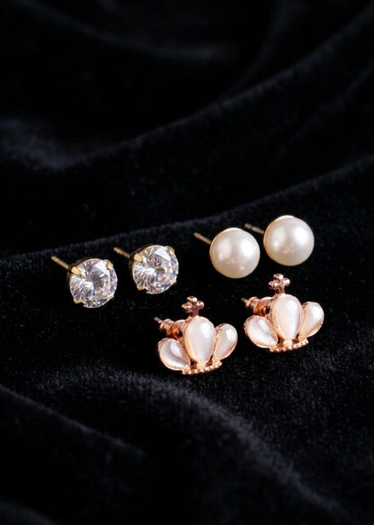 Three pairs of stud earrings displayed on a black velvet background: two pairs with clear round gemstones and one pair with white pearl-like inlays in a rose gold crown setting.