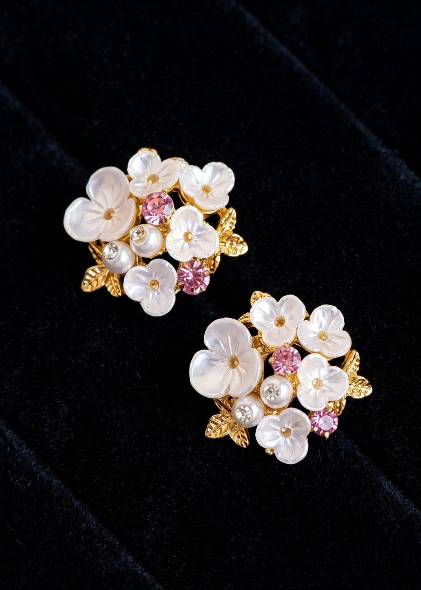 A pair of gold floral stud earrings featuring white mother-of-pearl flowers, small pearl-like beads, and pink crystal accents, displayed on a black velvet surface.