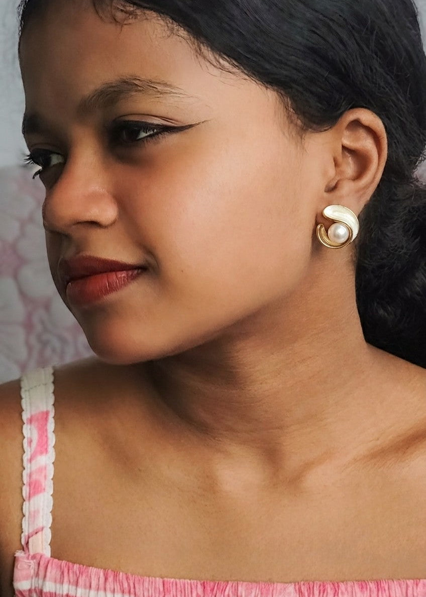A close-up side profile of a woman wearing a gold spiral-shaped stud earring with a white pearl accent, paired with a pink and white striped top.