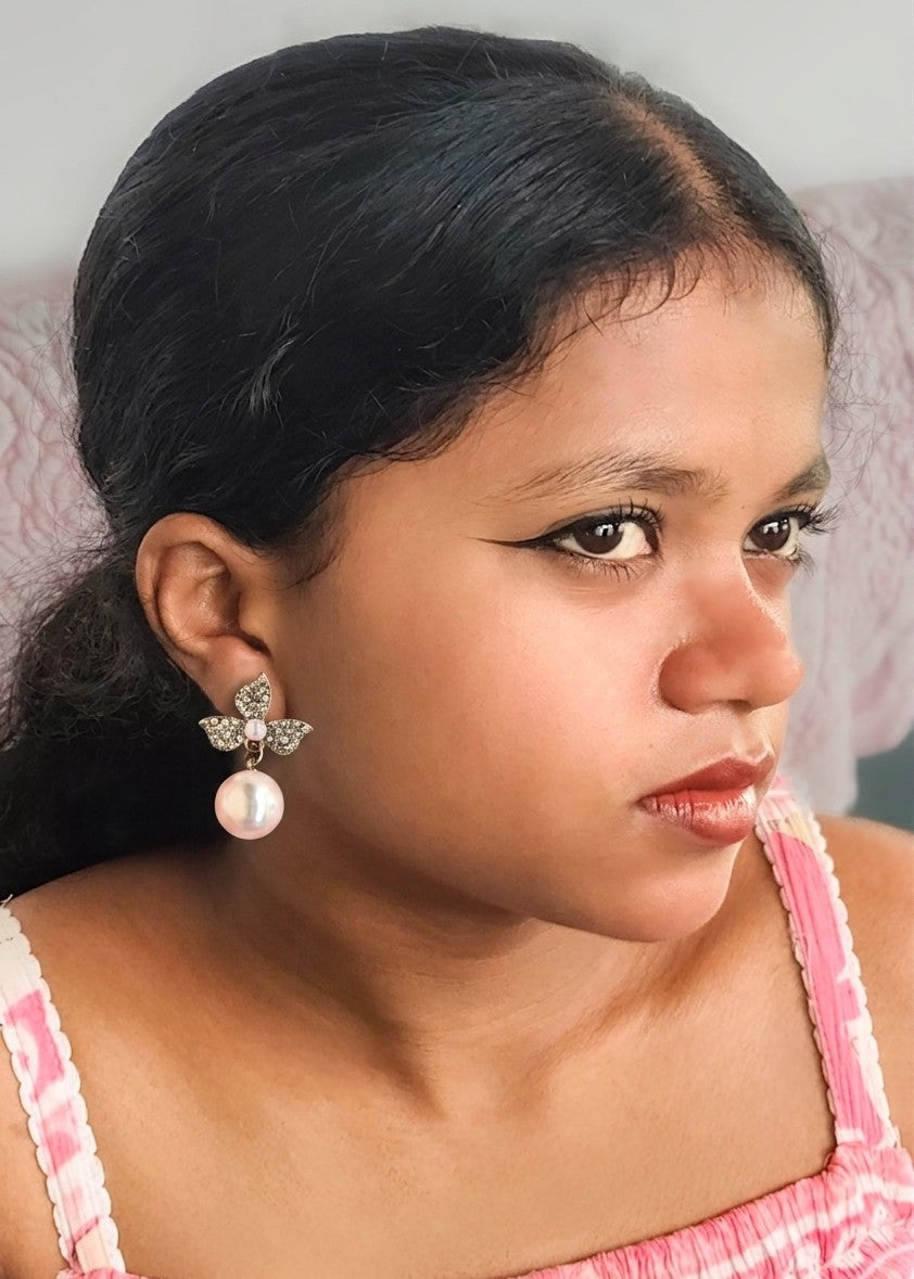 A close-up portrait of a young girl with dark hair, wearing a silver butterfly earring with a white pearl drop, and a pink striped top.