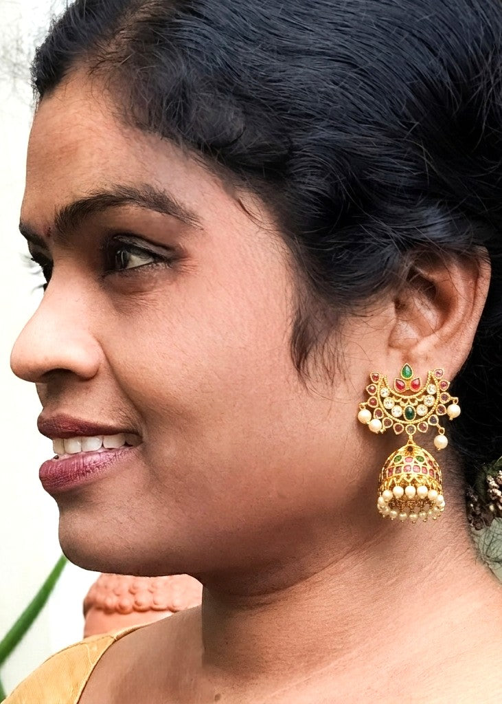 A close-up side profile of a woman wearing a traditional gold jhumka earring with green, red, and white stone accents and small pearl-like beads.