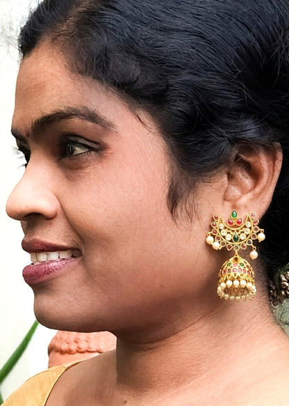 A close-up side profile of a woman wearing a traditional gold jhumka earring with green, red, and white stone accents and small pearl-like beads.
