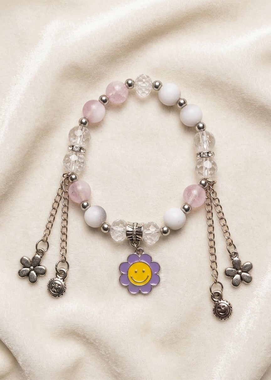 A beaded bracelet with pink and white beads, clear faceted beads, silver-toned chains, and a purple flower charm with a yellow smiley face, displayed on a cream fabric background.