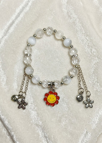 A beaded charm bracelet featuring white and clear faceted beads, silver-toned metal chains, and three dangling charms: a red flower with a yellow smiley face, a silver flower, and a silver star, displayed on a textured cream fabric background.