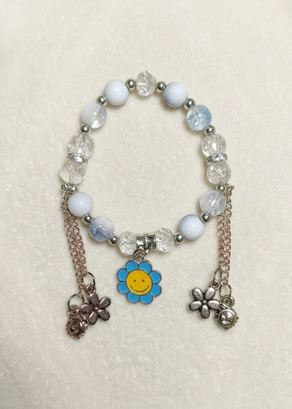 A beaded stretch bracelet with white and light blue beads, faceted clear crystal beads, silver-toned spacers, and three dangling charms: a blue flower with a yellow smiley face, a rose-gold-toned flower, and a silver-toned flower.