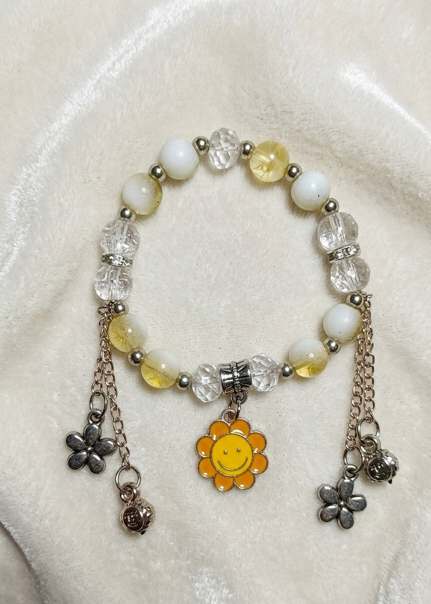 A beaded stretch bracelet featuring white and pale yellow beads, clear faceted beads, silver-toned metal spacers, and dangling charms including a yellow smiley face flower, a silver flower, and a small silver bell, displayed on a cream-colored fabric background.