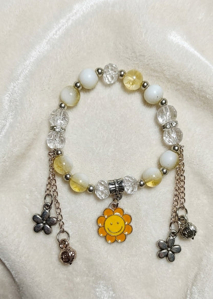 A beaded stretch bracelet featuring white and pale yellow beads, clear faceted beads, silver-toned metal spacers, and dangling charms including a yellow smiley face flower, a silver flower, and a small silver bell, displayed on a cream-colored fabric background.