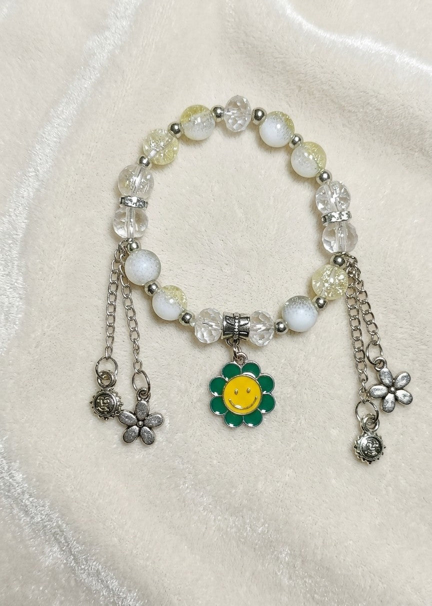 A beaded charm bracelet with white and pale yellow beads, silver-toned metal chains, and a central green and yellow flower charm, displayed on a soft cream-colored fabric background.
